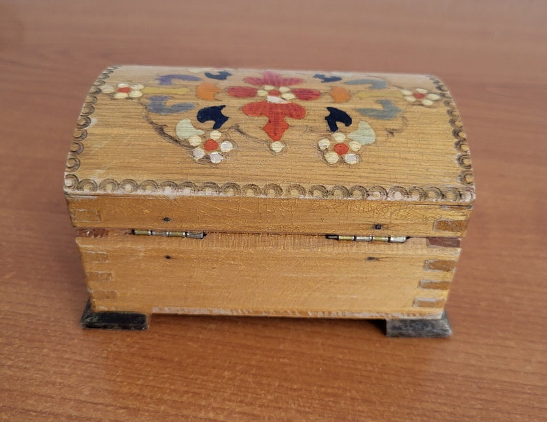 May include: A wooden box with a hinged lid. The lid is decorated with a colourful floral design painted in red, yellow, blue, and green. The box has a dark brown base.