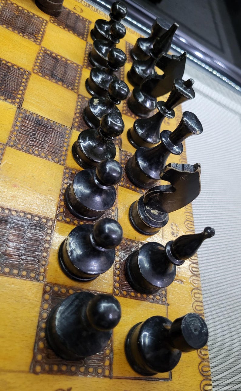 May include: A close-up of a chessboard with black chess pieces. The pieces are arranged in a row on the board, with the pawns in the front and the other pieces behind them. The board is made of wood and has a light brown color.