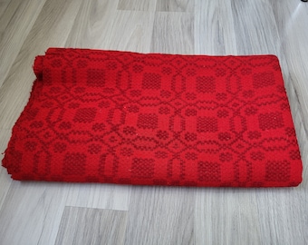 Peut inclure: Une couverture tissée rouge avec un motif géométrique. La couverture est pliée en deux et posée sur un sol en bois.