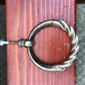 Può includere: Una maniglia ad anello in metallo argentato con un design attorcigliato. La maniglia è fissata a una superficie in legno con una vite.
