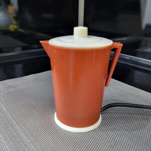 Può includere: Una caffettiera elettrica marrone con un coperchio bianco e una base bianca. La caffettiera ha un manico sul lato e un cavo che esce dalla base.