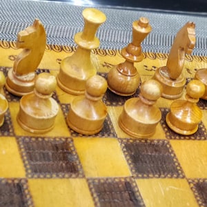 May include: A wooden chess set with light brown pieces on a brown and white checkered board. The pieces are arranged in a starting position for a game of chess.