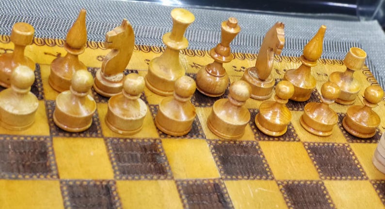 May include: A wooden chess set with light brown pieces on a brown and white checkered board. The pieces are arranged in a starting position for a game of chess.
