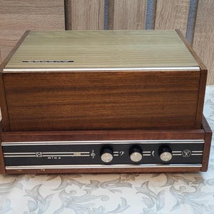 May include: A vintage wooden record player with a brown wood cabinet and a black control panel with knobs and the word "RIGA" printed on it.