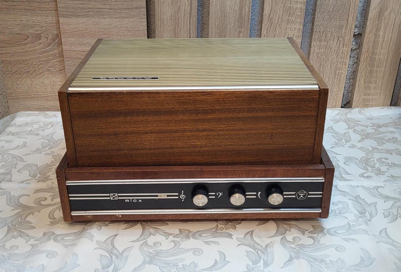 May include: A vintage wooden record player with a brown wood cabinet and a black control panel with knobs and the word "RIGA" printed on it.