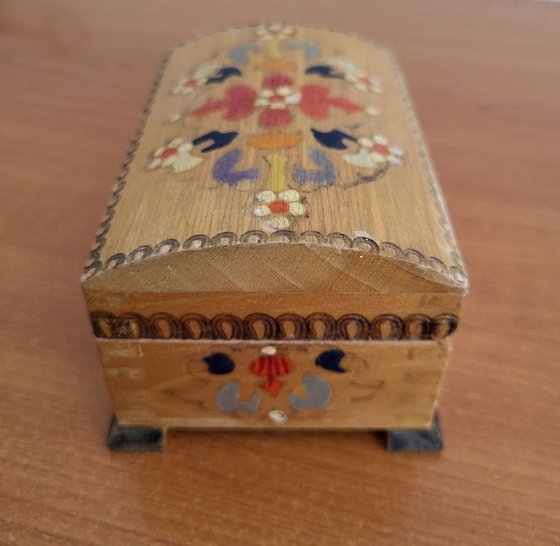 May include: A small wooden box with a hinged lid. The box is decorated with colourful painted flowers and a geometric pattern. The box has a dark brown trim around the edges.