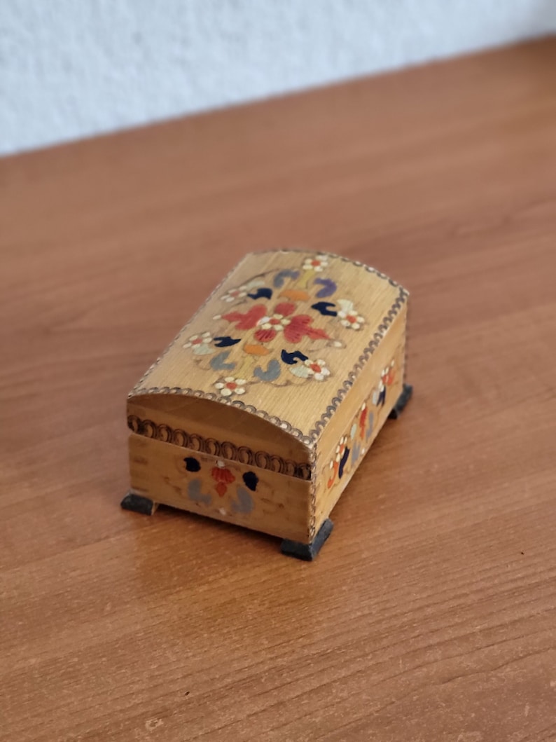 May include: A small wooden box with a hinged lid and painted floral designs in shades of red, blue, yellow, and green. The box has four small feet.