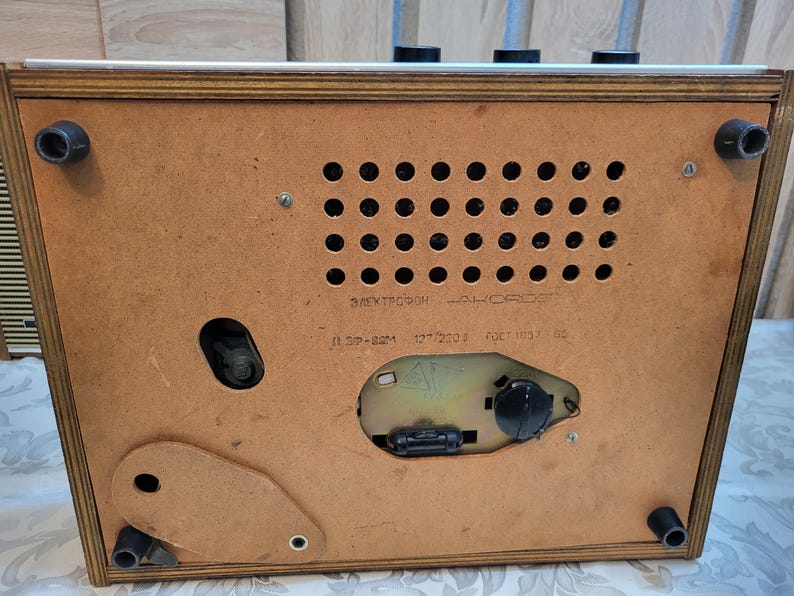 May include: A vintage wooden record player with a brown wood case and a metal control panel. The back of the player has a label that reads "Электрофон "АККОРД" 1.5ГР-82М 127/220В ФАСТ 1137 82."