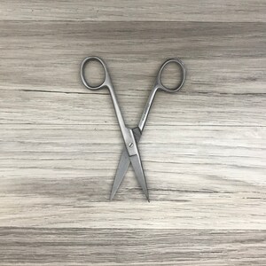 May include: A pair of stainless steel surgical scissors with a closed blade. The scissors are lying on a light brown wooden surface.