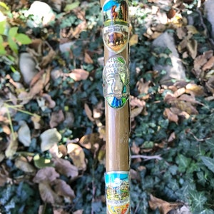 Puede incluir: Un bast&oacute;n de senderismo vintage hecho de bamb&uacute; marr&oacute;n claro, adornado con varias insignias met&aacute;licas. Las insignias presentan im&aacute;genes coloridas de paisajes, animales y texto, incluyendo "Wiedeburge", "Auerichtsplatz" y "Vogelpark Walsrode".