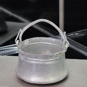 May include: A silver pot with a handle, the pot is sitting on a gray textured surface.