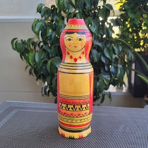 May include: A hand-painted wooden doll with a red, yellow and green design. The doll has a face with black eyes, a red mouth and a red necklace. The doll is wearing a yellow dress with a red skirt and a green and yellow patterned bottom.