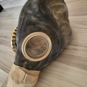 May include: A black rubber gas mask with a gold-colored metal valve and a clear plastic eye piece. The mask is covered in dirt and dust.