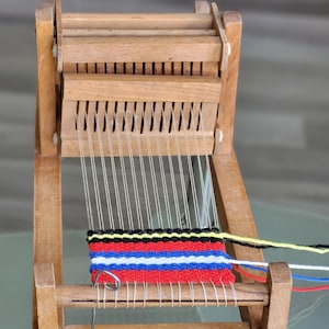 以下が含まれることがあります： 部分的に完成した織り部分のある小さな木製の織機。織られた部分は、赤、青、白、黒、黄色の糸が特徴です。織機は明るい色の木材で作られており、シンプルなデザインです。