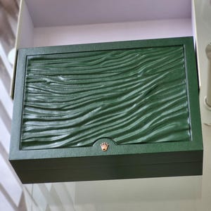 Può includere: Una scatola verde in pelle con un logo a corona dorata. La scatola presenta un motivo testurizzato a forma di onda.