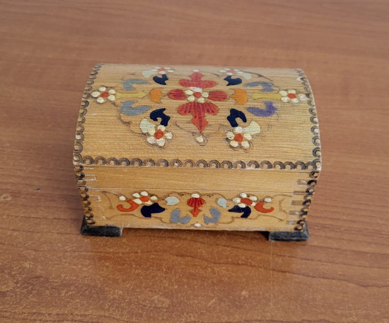 May include: A small wooden box with a hinged lid. The box is painted with a floral design in shades of red, orange, yellow, blue, and green. The box has a scalloped edge.