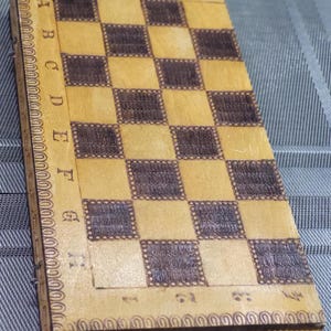 May include: A wooden chessboard with a brown and beige checkered pattern. The board is surrounded by a decorative border with a floral design.