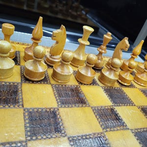 May include: A wooden chess set with light brown pieces on a light brown and dark brown chessboard. The pieces are arranged in a starting position for a game of chess.
