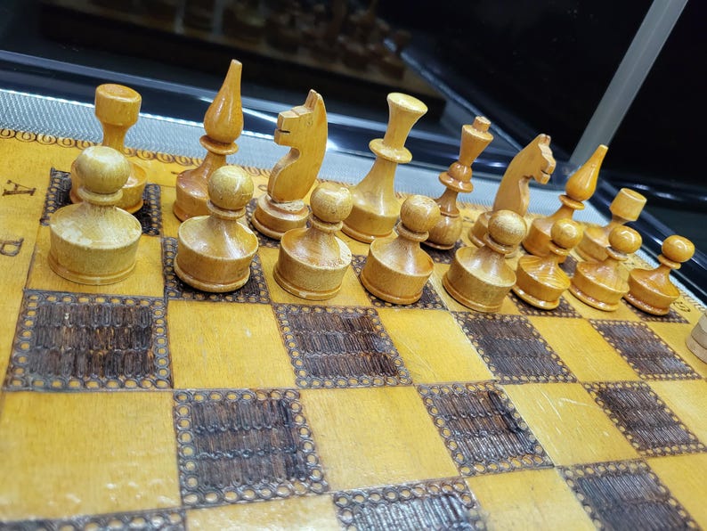May include: A wooden chess set with light brown pieces on a light brown and dark brown chessboard. The pieces are arranged in a starting position for a game of chess.
