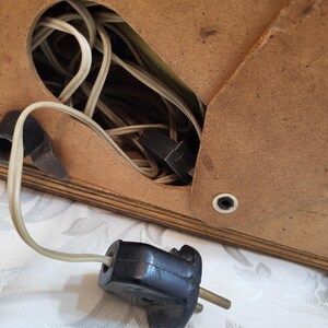 May include: A close-up of a black electrical plug with two prongs. The plug is connected to a white cord that is coiled inside a wooden box.