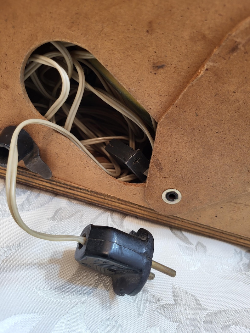 May include: A close-up of a black electrical plug with two prongs. The plug is connected to a white cord that is coiled inside a wooden box.