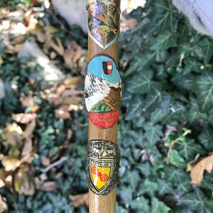 Puede incluir: Un bast&oacute;n de madera con varias insignias met&aacute;licas decorativas. Las insignias presentan escenas de monta&ntilde;a, texto y un dise&ntilde;o de escudo. El bast&oacute;n es de color marr&oacute;n claro y las insignias tienen varios colores, incluyendo azul, plata y amarillo.
