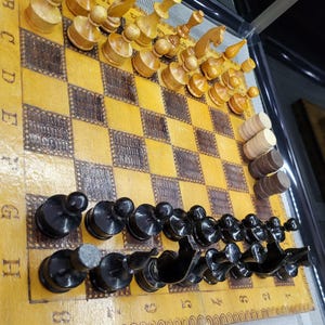 May include: A wooden chessboard with black and white squares. The board has a brown border with a decorative pattern. There are black and white chess pieces on the board. The board is in a glass case.