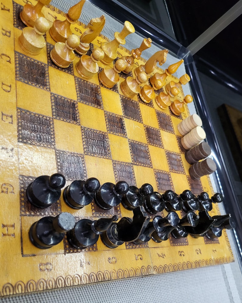 May include: A wooden chessboard with black and white squares. The board has a brown border with a decorative pattern. There are black and white chess pieces on the board. The board is in a glass case.