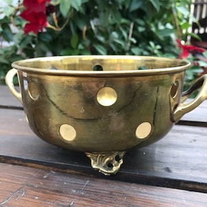 May include: A gold-coloured brass planter with a polka dot pattern and two handles. The planter has three feet and is sitting on a wooden surface.