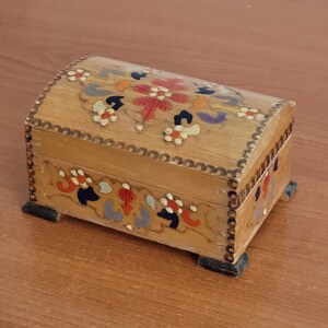 May include: A small wooden box with a hinged lid. The box is painted with a floral design in shades of red, orange, yellow, blue, and green. The box has a scalloped edge and is decorated with a beaded trim.