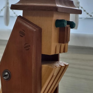 Peut inclure: Sculpture en bois représentant une figure avec un chapeau, des yeux verts et un design géométrique. La sculpture est faite de bois brun clair et foncé, avec des assemblages visibles et une vis métallique. La figure a un visage stylisé.