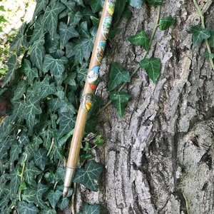Puede incluir: Un bast&oacute;n de madera vintage con varias insignias de recuerdo adjuntas. El bast&oacute;n marr&oacute;n claro tiene una punta plateada y se coloca contra una corteza de &aacute;rbol y hiedra verde. Las insignias parecen ser de varios lugares.