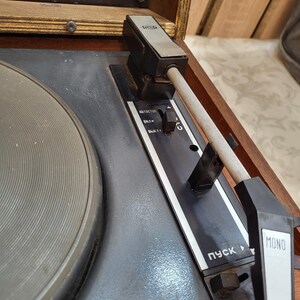 May include: Close-up of a vintage record player with a black and silver tone arm, a black turntable, and a wooden cabinet. The player has a black control panel with the words "AUTOSTOP", "ВКЛ/", "ВЫКЛ/" and "ПУСК" in Cyrillic script. The player also has a black and white "MONO" button.