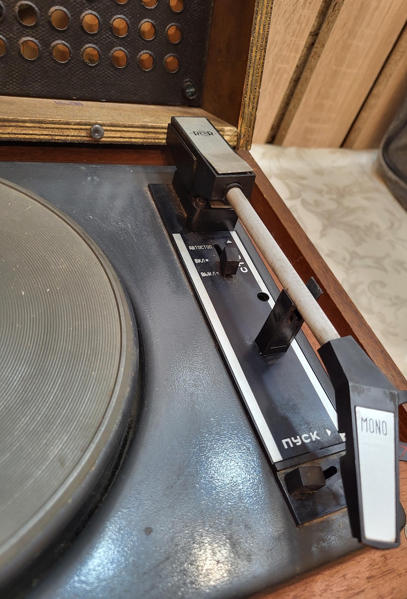 May include: Close-up of a vintage record player with a black and silver tone arm, a black turntable, and a wooden cabinet. The player has a black control panel with the words "AUTOSTOP", "ВКЛ/", "ВЫКЛ/" and "ПУСК" in Cyrillic script. The player also has a black and white "MONO" button.