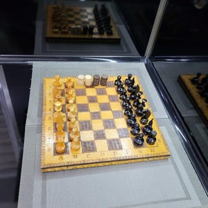 May include: A wooden chessboard with black and white squares. The board has a decorative border and the letters A through H and numbers 1 through 8 are printed on the sides. There are black and white chess pieces on the board.
