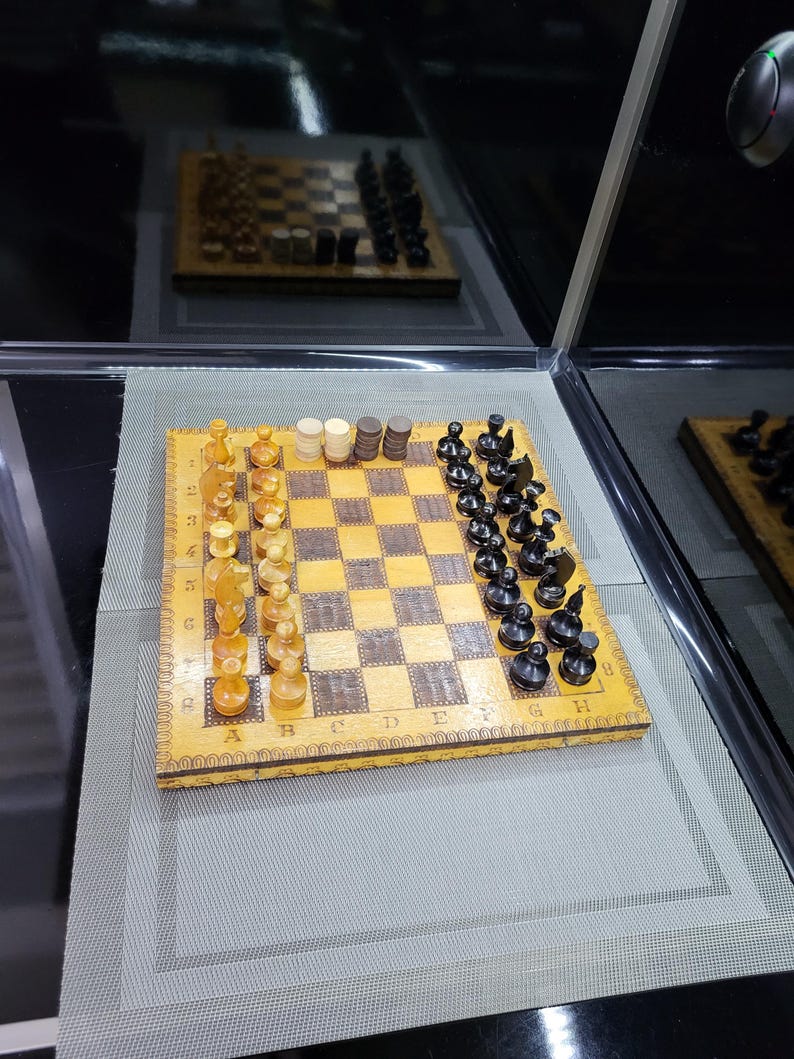May include: A wooden chessboard with black and white squares. The board has a decorative border and the letters A through H and numbers 1 through 8 are printed on the sides. There are black and white chess pieces on the board.