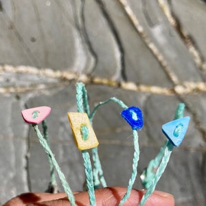 Pode incluir: Quatro botões de plástico coloridos presos a cordões turquesa. Os botões são rosa, amarelo, azul e azul claro.