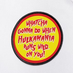 Könnte beinhalten: Weißer Rucksack mit einem kreisförmigen Aufnäher. Der Aufnäher zeigt den Text "Whatcha Gonna Do When Hulkamania Runs Wild On You!" in Rot und Gelb mit einem schwarzen Rand.