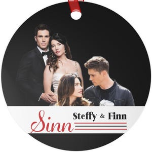 May include: A round, black Christmas ornament with a red ribbon. The ornament features a photo of four people. The text "Sinn Steffy & Finn" is printed in red and black on a white banner.