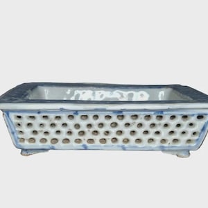 May include: Rectangular ceramic bonsai pot with a white and blue design. The pot has a perforated side pattern and a dark blue rim. It is supported by small feet and is suitable for small plants.