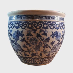 Puede incluir: Una maceta de cerámica azul y blanca con una forma ancha y redondeada. La maceta presenta un diseño detallado de dragones, flores y follaje. El borde tiene un patrón geométrico, y la estética general es tradicional y decorativa.