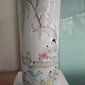 Può includere: Un vaso alto in ceramica bianca con una scena dipinta a mano di figure sotto un albero con fiori rosa. Il vaso ha una forma cilindrica e un bordo dorato. L'opera d'arte raffigura un ambiente esterno sereno.