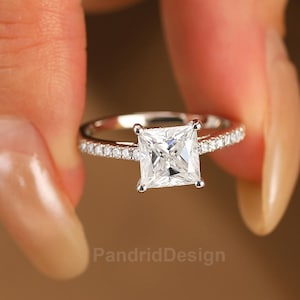 Könnte beinhalten: Ein silberner Ring mit einem quadratischen, klaren Edelstein. Das Band ist mit kleinen, klaren Edelsteinen verziert. Der Ring wird zwischen zwei Fingern vor einem verschwommenen Hintergrund gehalten. Der Text "PandridDesign" ist sichtbar.