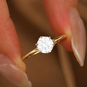 May include: A gold ring featuring a single round, brilliant-cut clear gemstone held in a six-prong setting. The band is smooth, polished, and slender, showcasing a classic solitaire design that highlights the clarity and sparkle of the center stone.