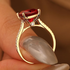 May include: A gold ring with a large, heart-shaped red gemstone as the centerpiece. The band is adorned with small, sparkling stones. A small, round diamond is set below the main stone. The ring is held up by a person's fingers.