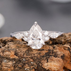 May include: A silver ring with a pear-shaped diamond center stone and smaller accent stones. The ring is set on a textured, brown wooden surface, with a blurred background of white and gray.