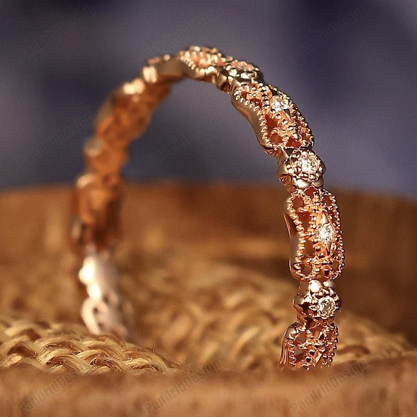 May include: A rose gold ring with a delicate filigree design and small, sparkling gemstones. The ring is set against a blurred background and rests on a wooden surface, showcasing its intricate details.