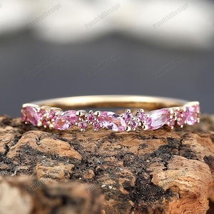 Könnte beinhalten: Ein zarter Goldring mit einer Reihe von rosafarbenen Marquise- und Rundsteinen. Der Ring liegt auf einer Holzoberfläche.