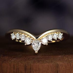 May include: A gold-colored ring with a curved design, set with multiple clear, sparkling gemstones. The central stone is pear-shaped, and the ring is displayed on a wooden surface, showcasing its elegant details.