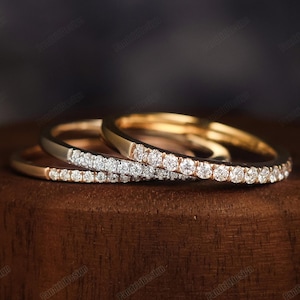 May include: Three gold rings with small, sparkling white gemstones set in a row. The rings are displayed on a dark wooden surface, with a blurred background. The rings are likely wedding bands or fashion jewelry.